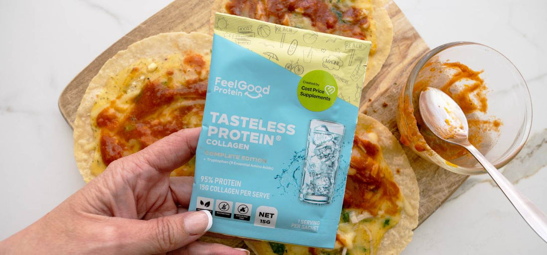 Top-down photo of a hand holding a Tasteless Protein Collagen by Feel Good Protein sachet over the centre of a wooden board with three pizzas, a small glass bowl, and spoon on a white surface.