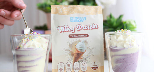 Front-facing photo of two No Bake High Protein Cheesecake Cups with a yellow and white sachet of Whey Protein by Feel Good Protein (Vanilla Ice Cream) in the centre, with a softly blurred modern kitchen in the background.
