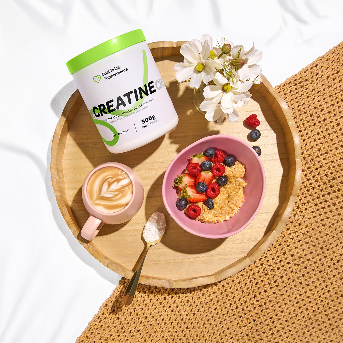 Wooden tray with creatine supplement container, bowl of berries, coffee, and flowers on a white background