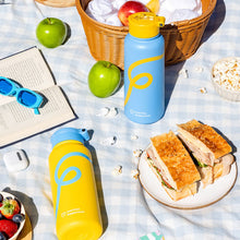 Colourful Blue and Yellow water bottles on a picnic blanket surrounded by apples, picnic basket and sandwich