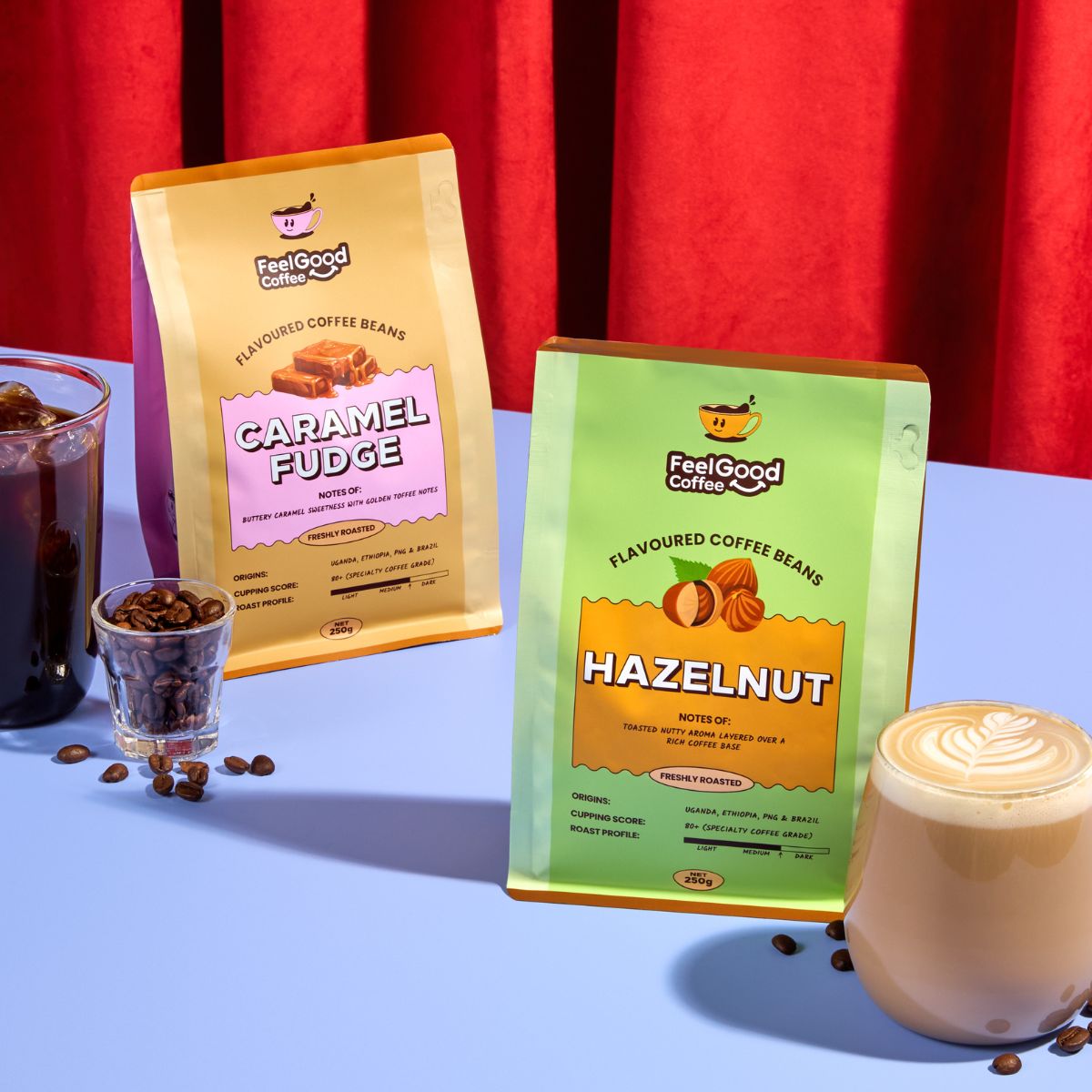 Two packages of FeelGood Coffee Flavoured Coffee Beans with a glass of coffee and a cup of cappuccino on a blue surface with red curtains in the background.