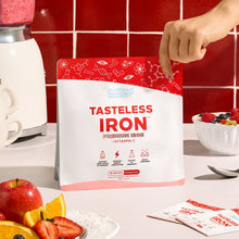 Tasteless Iron by Feel Good Protein in front of red tiled wall and next to fruit bowl