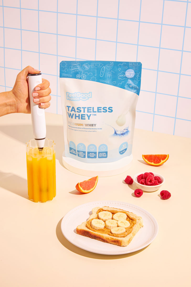 Person holding a bottle next to a bag of Tasteless Whey protein powder, with a plate of toast and fruit on a checkered background.